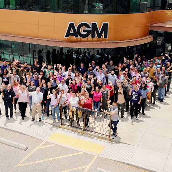 A large group of AGM employee's wave to the camera while standing outside the AGM building